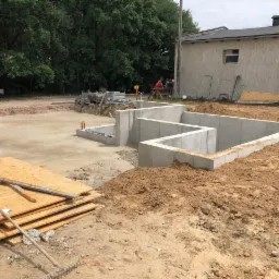 Wylany fundament pod budynek z widocznymi betonowymi ścianami, stos płyt drewnianych na pierwszym planie, narzędzia i grupa robotników w tle.