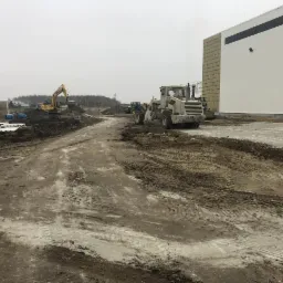 Teren budowy z widocznymi maszynami: równiarka, koparka i ciągnik, w tle hala magazynowa z jasną elewacją.