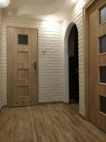 Korytarz po remoncie: ściana z białej cegły dekoracyjnej, drewniana podłoga panelowa i dwie drewniane drzwi z przeszkleniami, jedna z nich prowadzi do łukowatego przejścia.