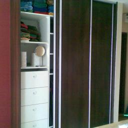 Stolpar - Wnętrze szafy przesuwnej z ciemnymi drzwiami i białymi elementami wewnętrznymi, widoczne półki z ręcznikami, szuflady i kosz z siatki.