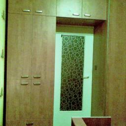 Stolpar - Wysoka szafa z płyty imitującej drewno, z górnymi szafkami i dolnymi szufladami, obok drzwi z szybą o wzorze z okręgów.