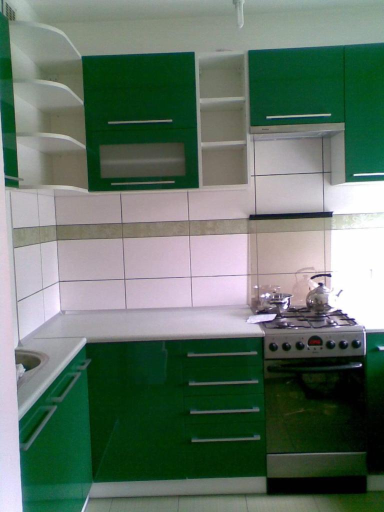 Zielona zabudowa kuchenna z białymi półkami i blatem, widoczna płyta gazowa z czajnikiem i garnkiem, białe płytki na ścianie z zielonym paskiem.