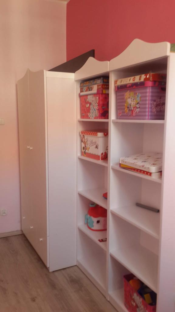 Biała szafa z otwartymi półkami w pokoju dziecięcym w Poznaniu, wypełniona pudełkami z motywami bajkowymi i zabawkami, pod różową ścianą.