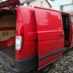 Czerwony bus z otwartymi tylnymi drzwiami, widoczna pusta przestrzeń ładunkowa z drewnianą podłogą i kratkami wentylacyjnymi, zaparkowany na kamienistym podłożu.