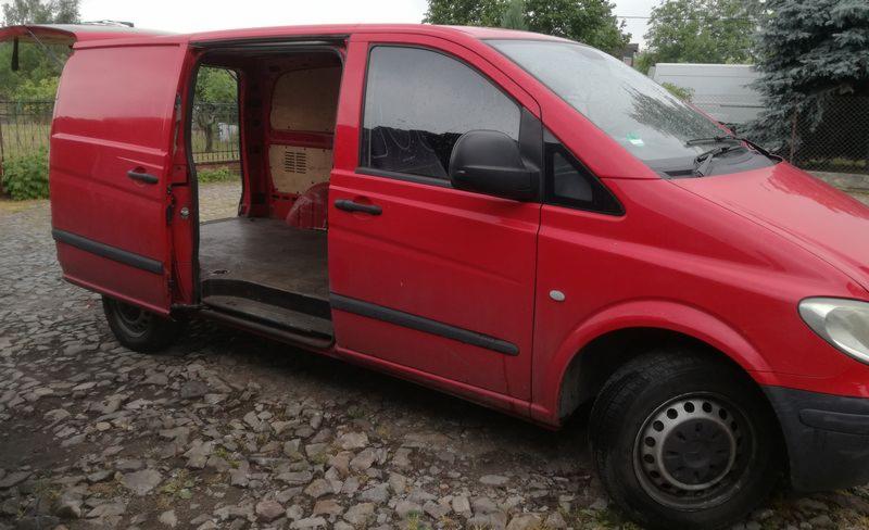 Czerwony bus z otwartymi drzwiami bocznymi i tylnymi, zaparkowany na żwirowym podłożu, widoczne wnętrze przestrzeni ładunkowej wyłożone płytami drewnianymi.