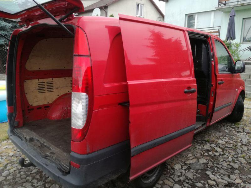 Czerwony bus z otwartymi tylnymi drzwiami, widoczna pusta przestrzeń ładunkowa z drewnianą podłogą i kratkami wentylacyjnymi, zaparkowany na kamienistym podłożu.