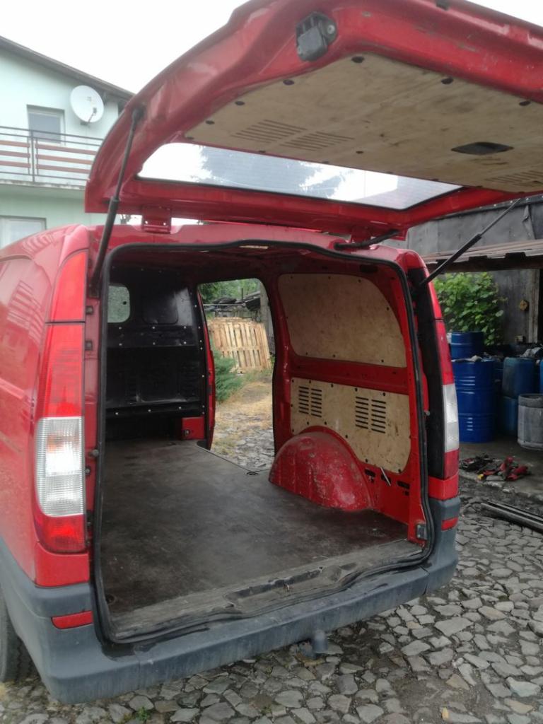 Czerwony bus z otwartymi drzwiami bagażnika, widoczna pusta przestrzeń ładunkowa z drewnianą podłogą, w tle palety i niebieskie beczki.