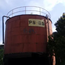 Zardzewiały, cylindryczny zbiornik przemysłowy z napisem PS-05, otoczony zielenią, z widoczną konstrukcją schodów i rur na tle błękitnego nieba.