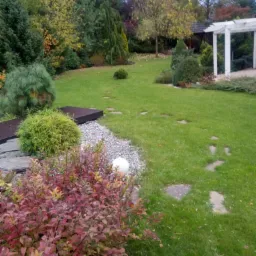 Jesienny ogród z drewnianym mostkiem, ścieżką z kamiennych płyt i białą kulą dekoracyjną na tle zielonego trawnika, drzew i białej altany.