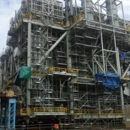 Rozbudowana konstrukcja przemysłowa w trakcie budowy, otoczona rusztowaniami i zabezpieczona plandekami, widok z poziomu gruntu.