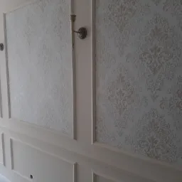 Ściana z dekoracyjną, jasną tapetą z motywem roślinnym, obramowana białymi listwami i kinkietami, widoczne niedokończone prace wykończeniowe.