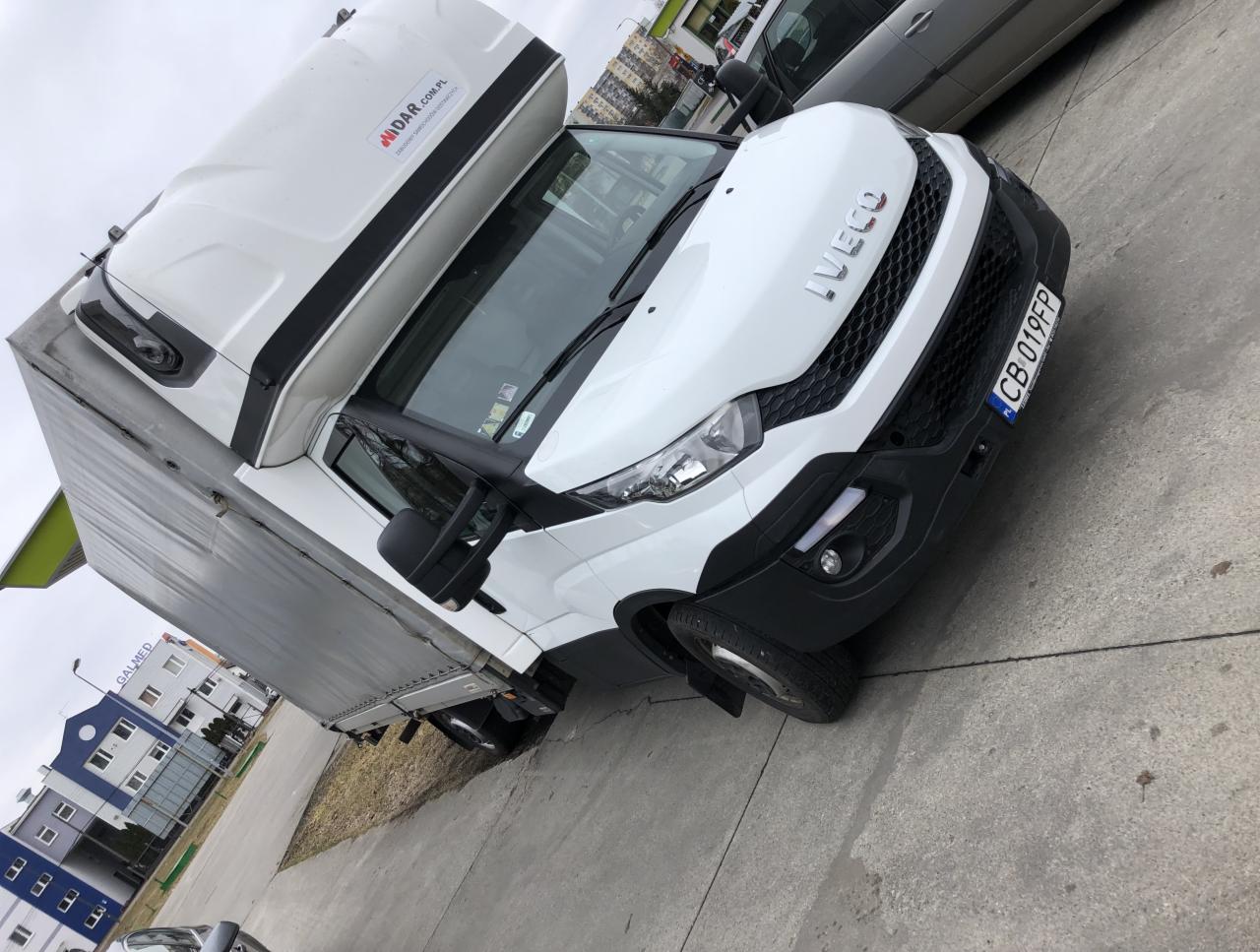 Biały samochód dostawczy Iveco z zabudową kontenerową i logo MIDAR.COM.PL, zaparkowany na betonowym placu, widoczny fragment budynku GALMED w tle.
