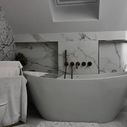 PLK STUDIO Spółka z o. o. - Nowoczesna łazienka z wolnostojącą białą wanną, złotą armaturą i marmurowymi płytkami na ścianach, uzupełniona o białe ręczniki na złotym stojaku i rośliny doniczkowe.