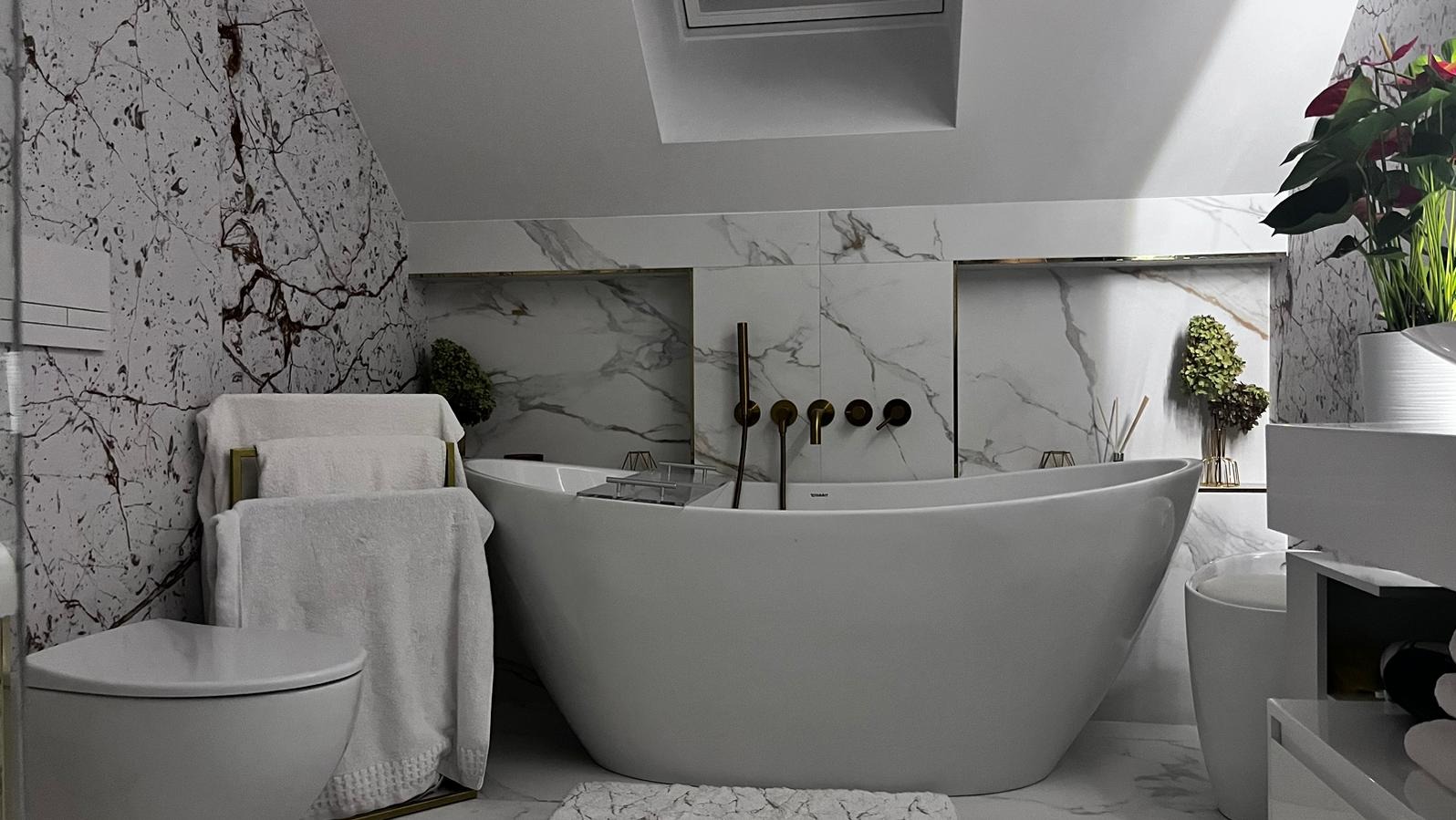 Nowoczesna łazienka z wolnostojącą białą wanną, złotą armaturą i marmurowymi płytkami na ścianach, uzupełniona o białe ręczniki na złotym stojaku i rośliny doniczkowe.