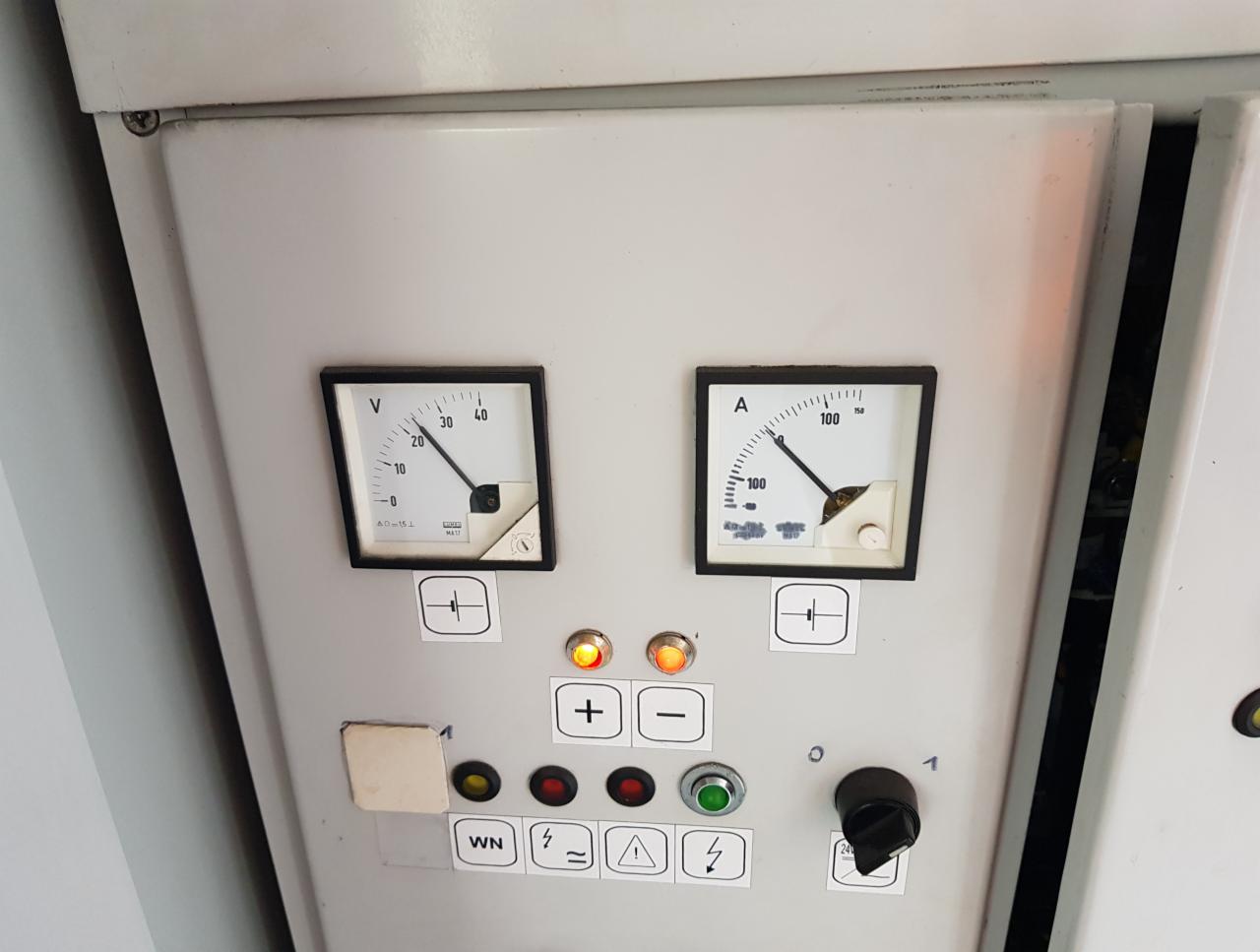 Panel sterowania z dwoma analogowymi miernikami wskazującymi napięcie i prąd, otoczony przyciskami i przełącznikami, widoczne oznaczenia symboli elektrycznych.