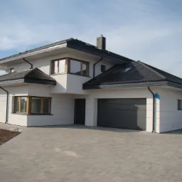 Nowoczesny dom jednorodzinny z szarą bramą garażową segmentową i jasną elewacją, widok z zewnątrz na podjazd wyłożony kostką brukową.