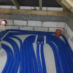 Niebieskie, karbowane rury wentylacyjne ułożone równolegle na podłodze w surowym pomieszczeniu z betonowymi ścianami i drewnianą konstrukcją dachu.