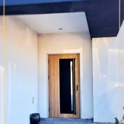 Nowoczesne drzwi wejściowe z drewnianą okładziną i czarnym, pionowym panelem szklanym, wkomponowane w jasną elewację budynku z ciemnym zadaszeniem.