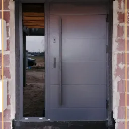 Szare drzwi wejściowe z bocznym przeszkleniem, zamontowane w surowym murze z widoczną izolacją i elementami montażowymi.