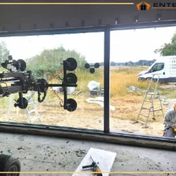 Montaż panoramicznego okna z czarną ramą za pomocą specjalistycznego sprzętu z przyssawkami, widoczny pracownik i firmowy bus w tle.