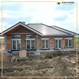 Budowa domu jednorodzinnego w trakcie montażu białych okien PCV z roletami zewnętrznymi, widoczne cegły elewacyjne, fundament i wykop pod budynek.