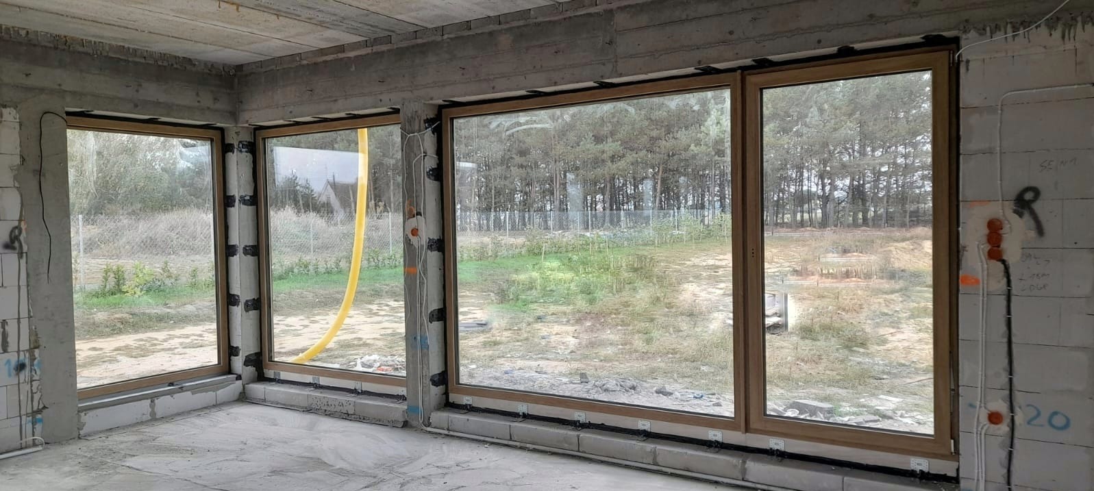 Montaż okien z drewnianą ramą w surowym stanie, widoczne przewody elektryczne i żółty wąż wentylacyjny.