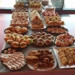 Bogato zastawiony stół z różnorodnymi przekąskami cateringowymi, w tym mini-kanapeczki, ciasteczka, kulki mięsne i baklawa, na szarym obrusie, z czerwonym tłem w tle.