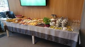 Stół przykryty białym obrusem z szarym wzorem, zastawiony różnorodnymi przekąskami, wędlinami, jajkami, ciastami, filiżankami i szklankami, z rośliną w doniczce pośrodku, na tle drewnianej ściany...