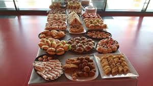 Bogato zastawiony stół z różnorodnymi przekąskami cateringowymi, w tym mini-kanapeczki, ciasteczka, kulki mięsne i baklawa, na szarym obrusie, z czerwonym tłem w tle.