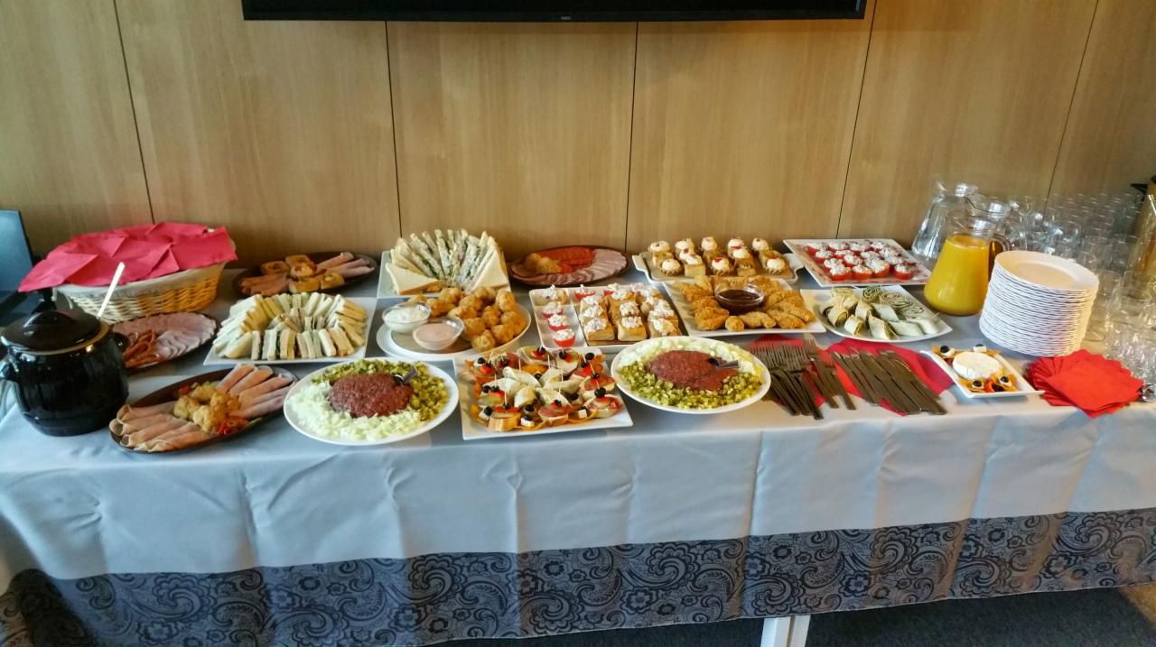 Stół z różnorodnymi przekąskami: kanapki, sałatki, roladki, mini desery, patery wędlin, sosy, dzbanek soku pomarańczowego, stos naczyń, sztućce, koszyk z serwetkami i garnkiem na tle drewnianej...