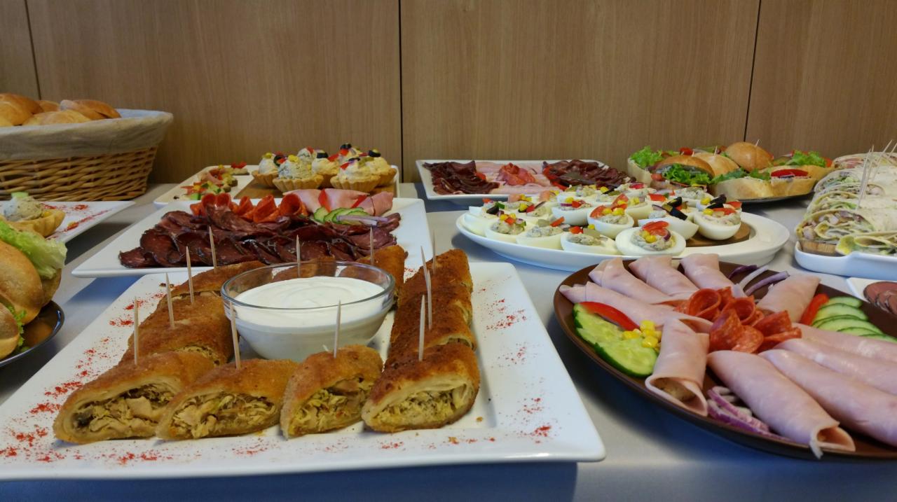 Bufet z różnorodnymi przekąskami: roladki z nadzieniem, jajka faszerowane, wędliny, kanapki i tartaletki, ułożone na białych talerzach na stole z jasnym tłem.