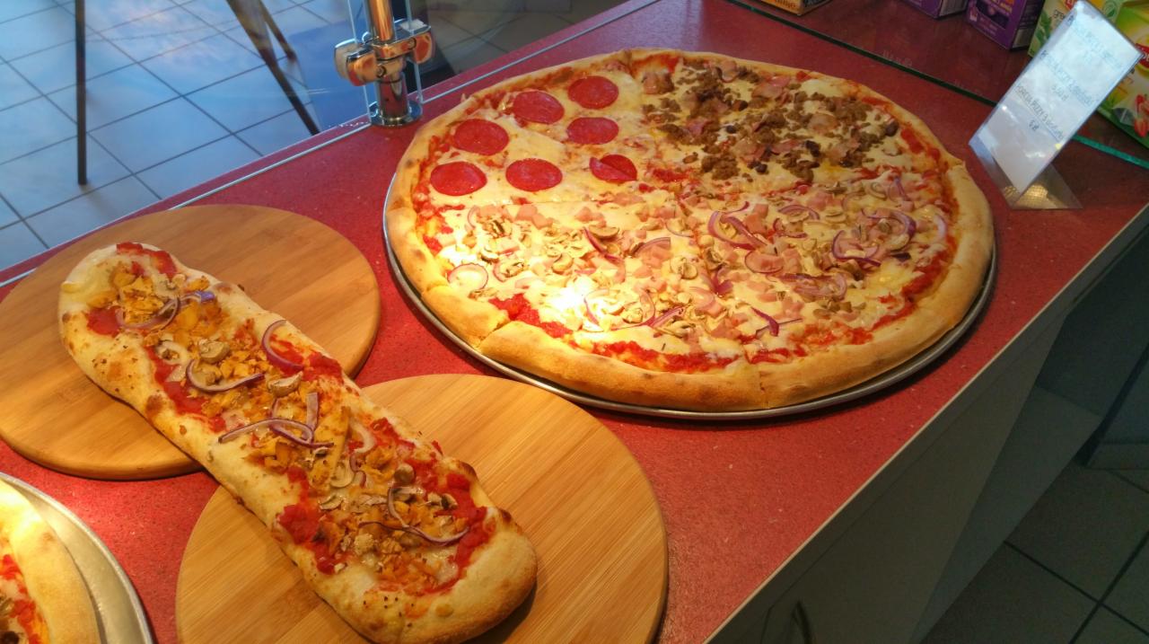 Duża pizza podzielona na cztery różne smaki: salami, mięsna, pieczarkowa i szynkowa z czerwoną cebulą, obok podłużna pizza na drewnianej desce.