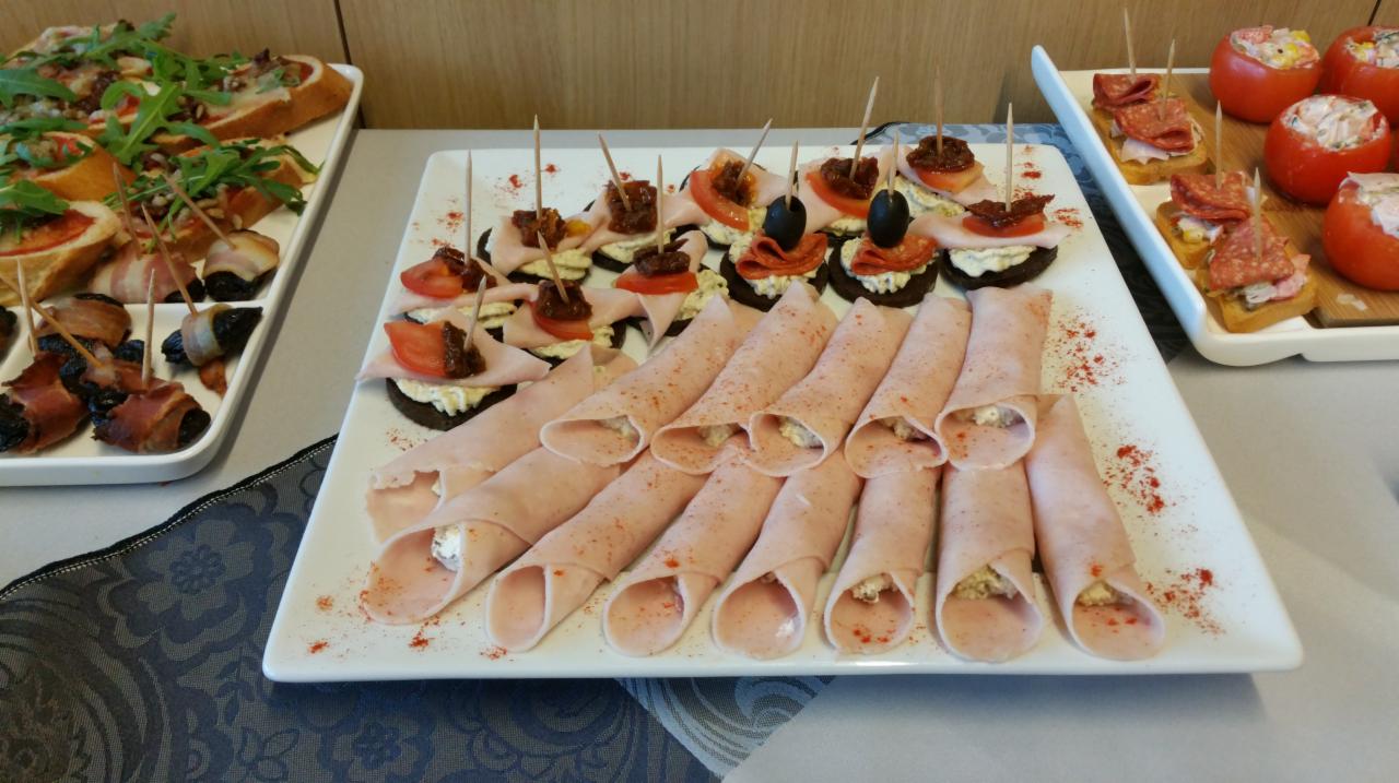 Dekoracyjnie ułożone przekąski na białych talerzach: roladki z szynki z kremowym nadzieniem posypane papryką, koreczki z suszonymi śliwkami owiniętymi boczkiem, mini kanapeczki z pastą, pomidorami...