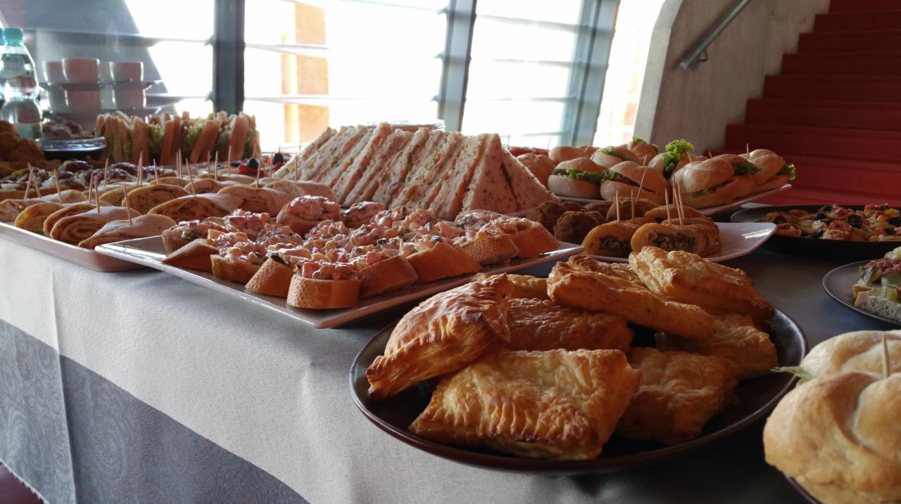 Stół z różnorodnymi przekąskami: kanapki trójkątne, tartinki z sałatką, mini burgery z sałatą, paszteciki z ciasta francuskiego, roladki i koreczki na paterach, w tle schody i okno.