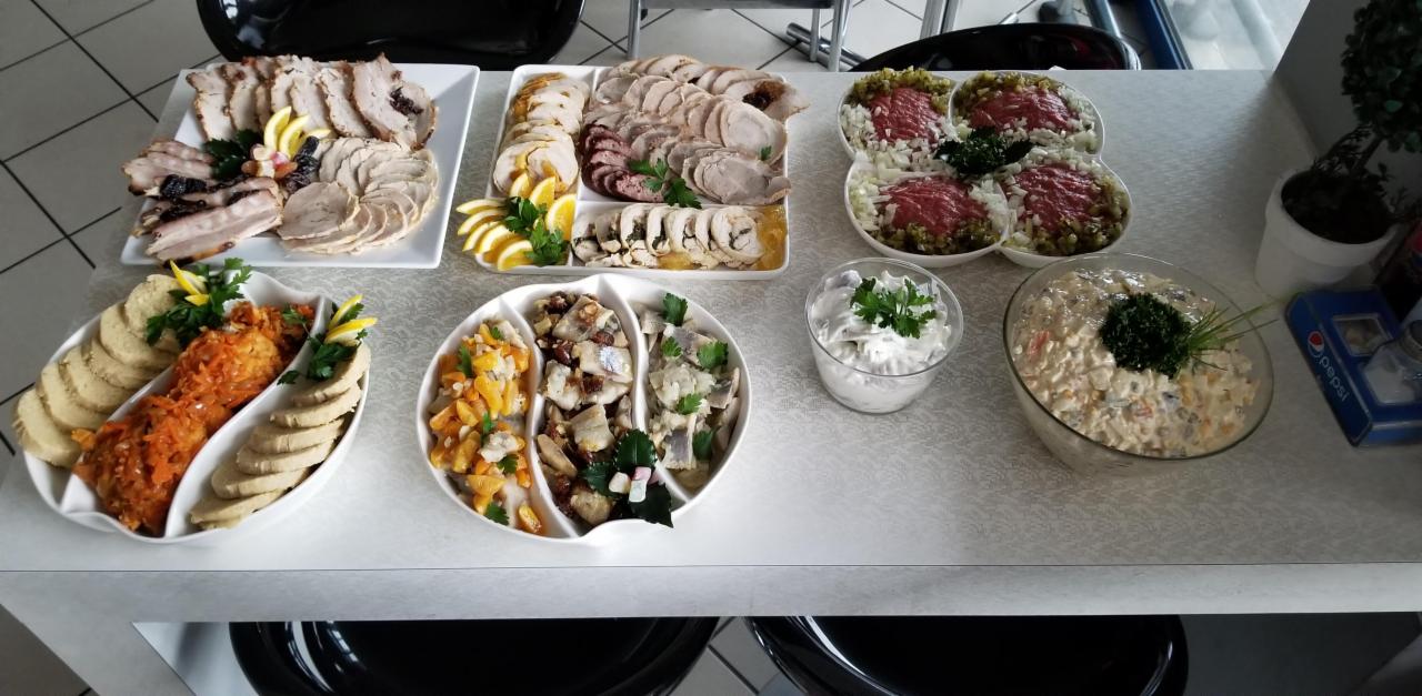 Bogato zastawiony stół z różnorodnymi potrawami: wędliny w plastrach dekorowane cytryną i ziołami, sałatki warzywne, tatar wołowy z cebulą i ogórkiem, kluski śląskie z sosem marchewkowym, śledzie...