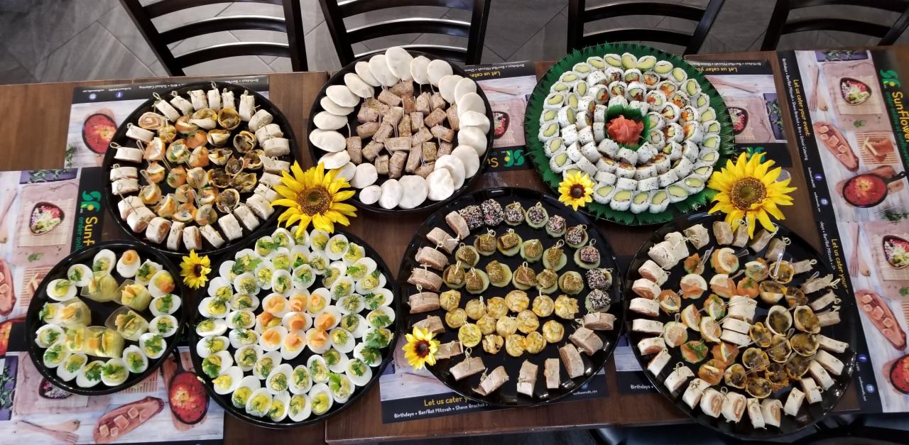 Różnorodny wybór przekąsek na ciemnych tacach, udekorowanych słonecznikami, ustawionych na stole z nadrukowanymi motywami kulinarnymi, widok z góry.