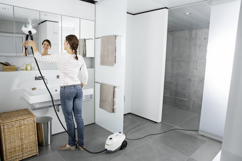 Kobieta w łazience czyści lustro parownicą, widoczny biały parownik na podłodze i para wydobywająca się z dyszy