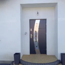 Nowoczesne wejście do domu z antracytowymi drzwiami z dekoracyjnym srebrnym panelem, jasna elewacja i kostka brukowa ułożona w półokrągły wzór.