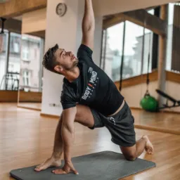 Mężczyzna w czarnej koszulce z logo 'BODY / MIND experts' wykonuje pozycję jogi na szarej macie w jasnym pomieszczeniu z drewnianą podłogą i lustrem.