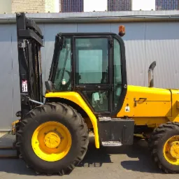 Żółty wózek widłowy z kabiną i dużymi oponami terenowymi, zaparkowany na zewnątrz budynku z szarej blachy falistej.