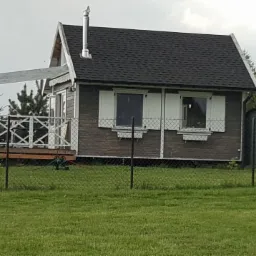 Niewielki, parterowy dom z ciemnego drewna z białymi okiennicami i zadaszonym tarasem, otoczony zielonym trawnikiem i ogrodzeniem z siatki.