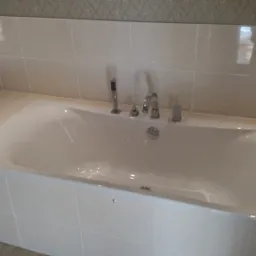 Wanna obudowana jasnymi, kwadratowymi płytkami ceramicznymi, z armaturą w kolorze chromu, wykończona listwą dekoracyjną na ścianie.