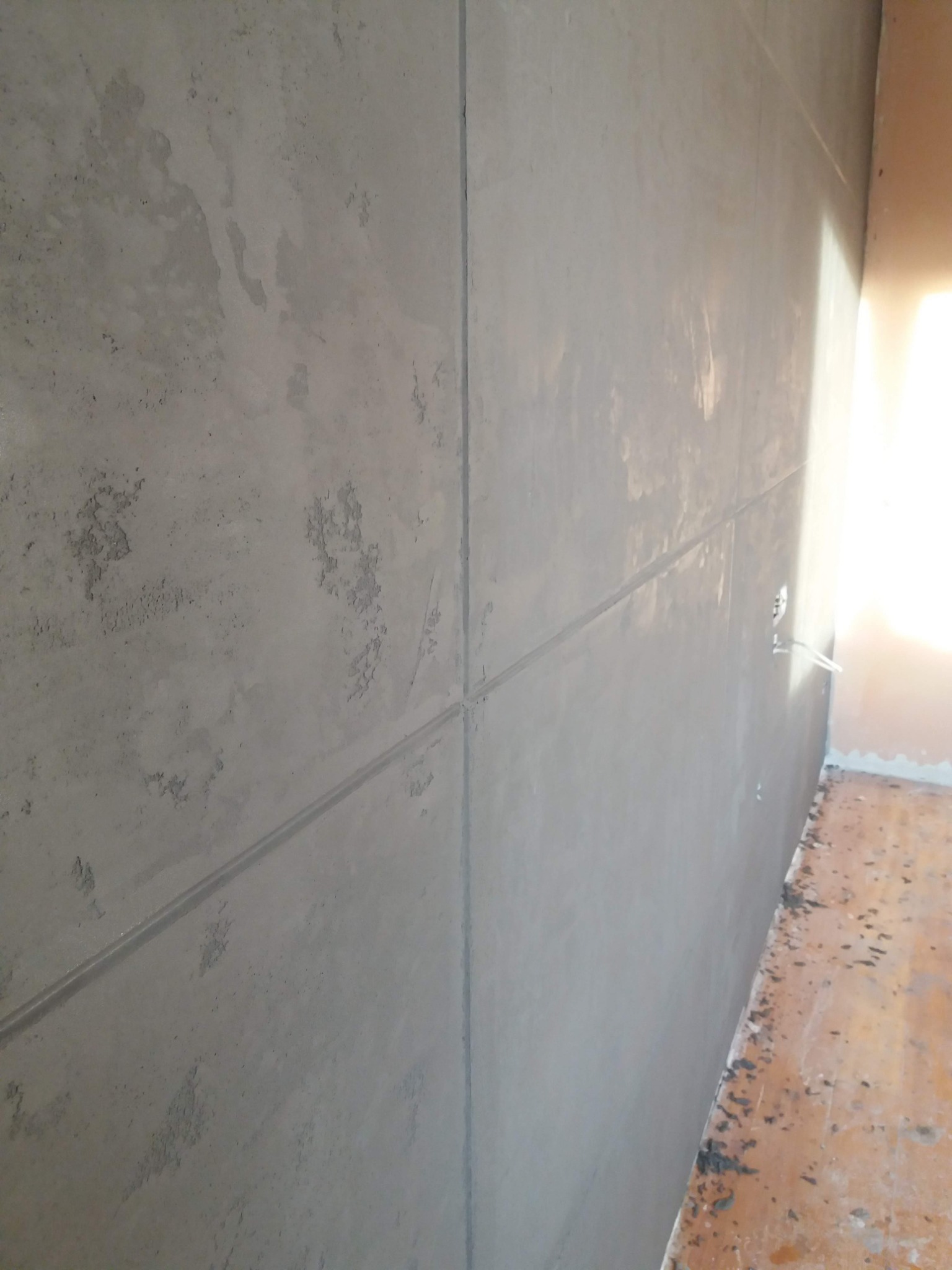 Ściana pokryta szarym betonem architektonicznym, podzielona na płyty fugami, widoczne gniazdko elektryczne i fragment podłogi w trakcie prac wykończeniowych.