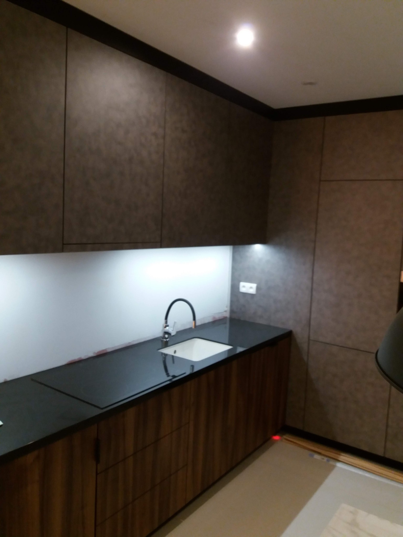 Nowoczesna kuchnia z ciemnymi, teksturowanymi szafkami górnymi i drewnopodobnymi szafkami dolnymi, czarnym blatem, białym fartuchem i minimalistyczną armaturą.