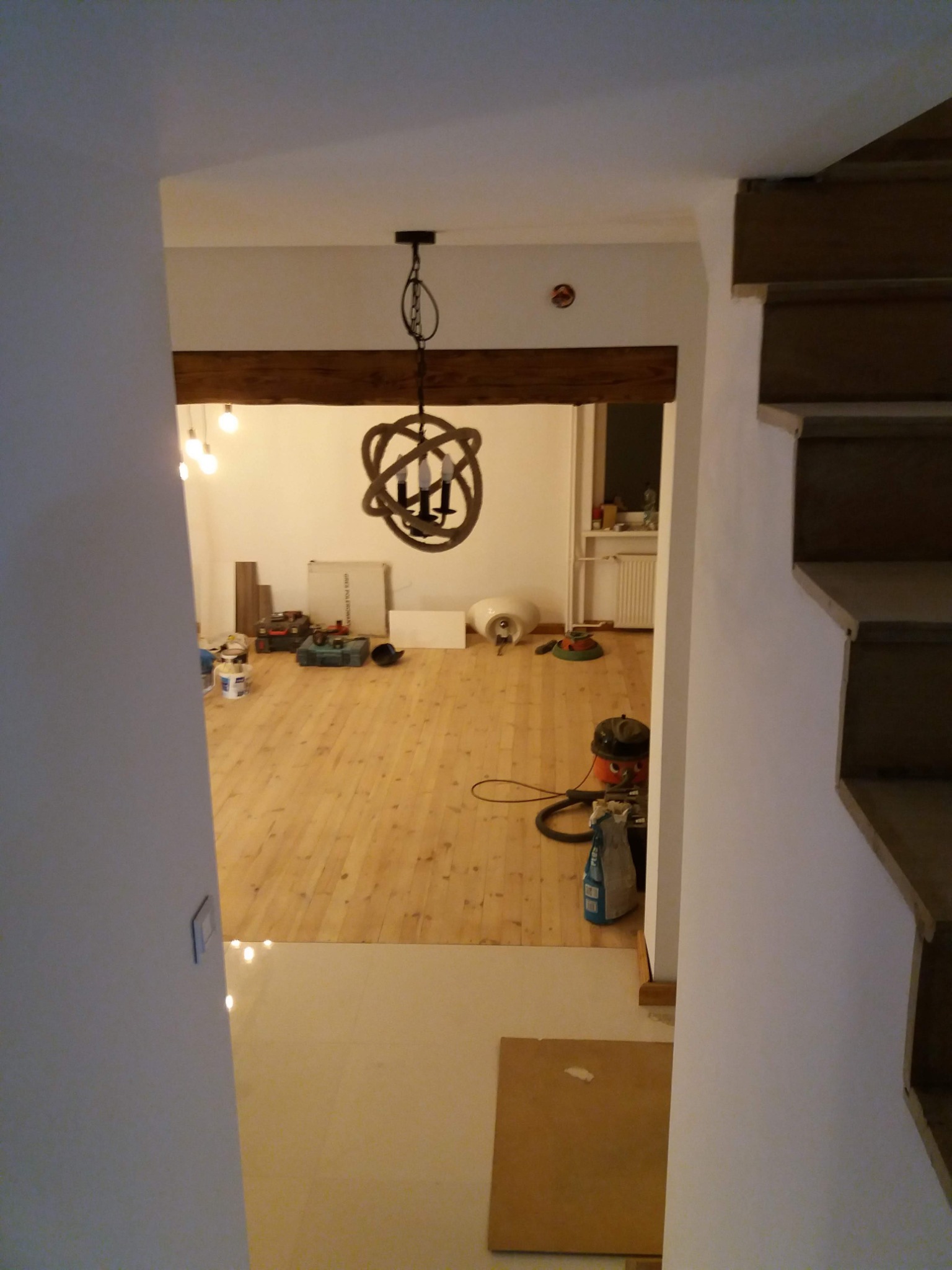 Widok na remontowane wnętrze z drewnianą podłogą i dekoracyjnym żyrandolem, widoczne narzędzia i materiały budowlane, ujęcie z klatki schodowej.