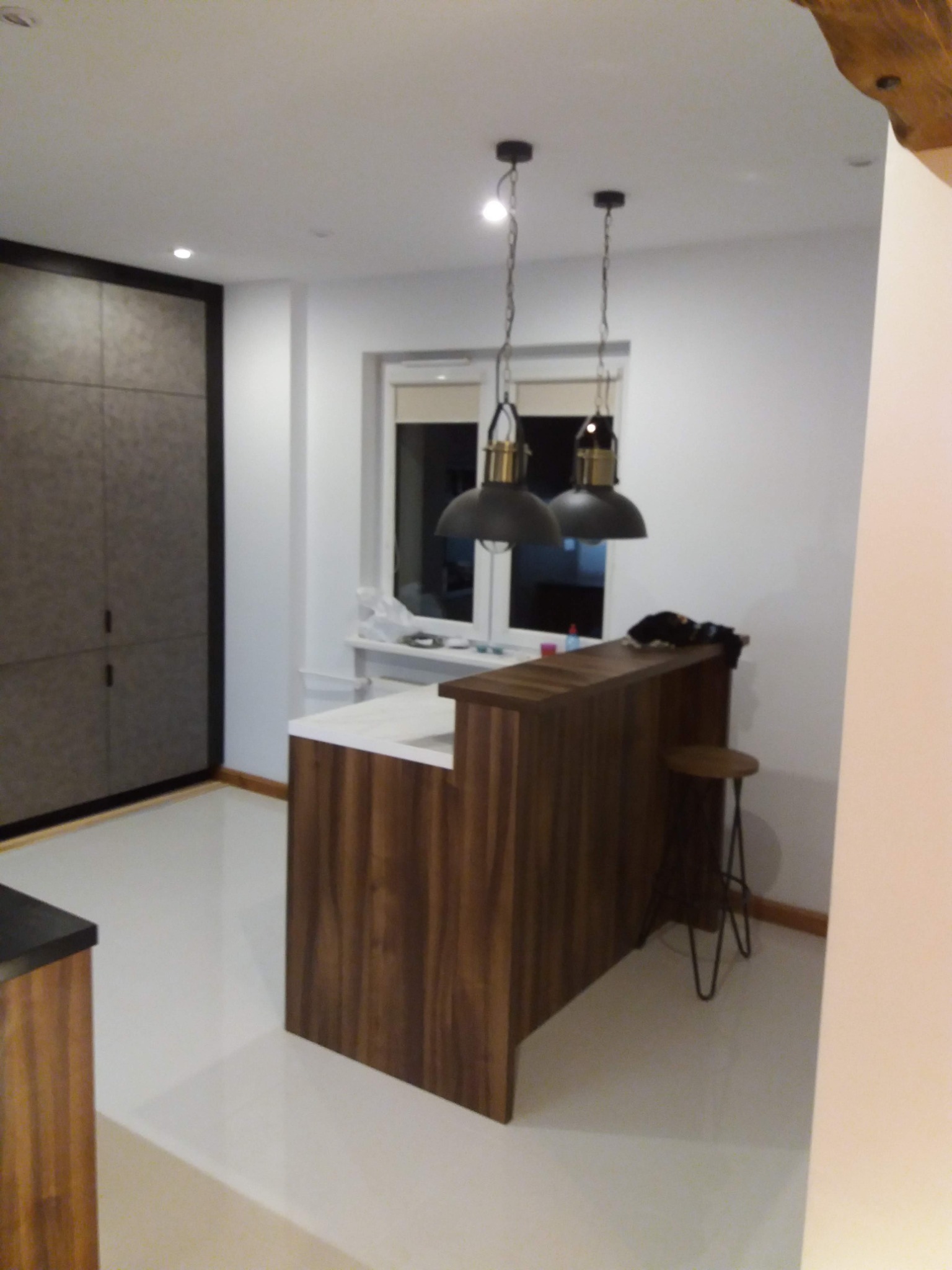 Nowoczesna kuchnia z drewnianą wyspą z białym blatem, oświetlona dwoma wiszącymi lampami w stylu industrialnym, widok na okno i minimalistyczną szafę w zabudowie.