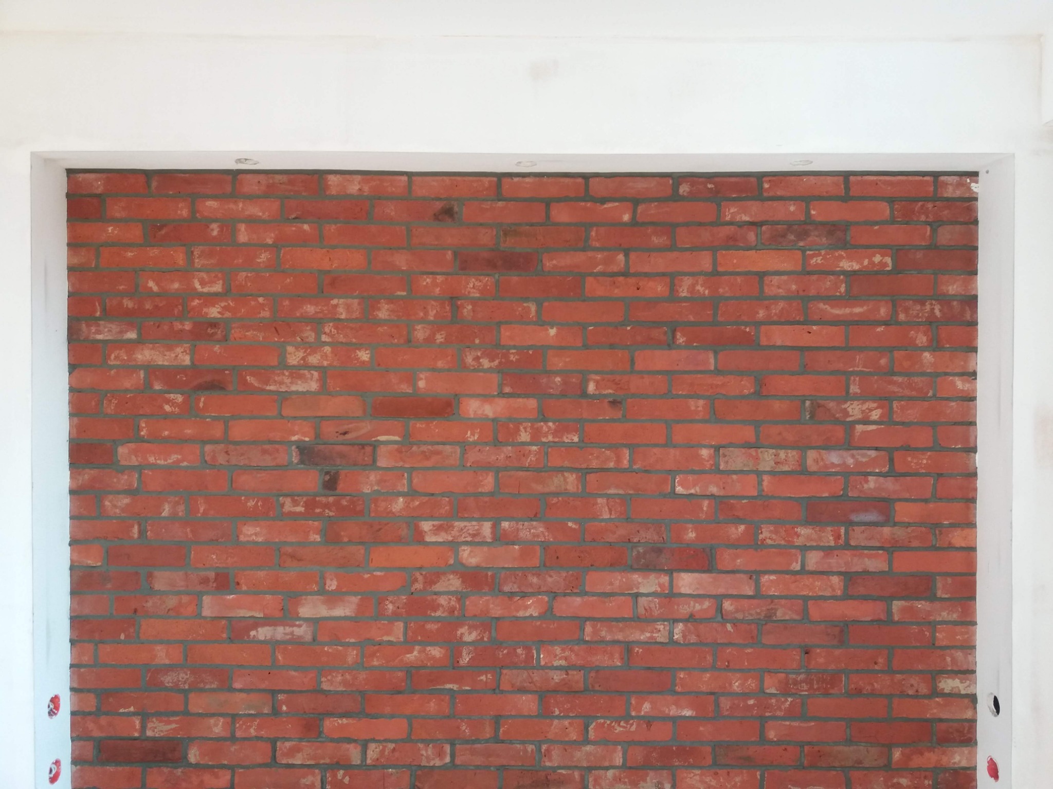 Czerwona ściana z cegły w białej ramie, widoczne punkty świetlne w suficie, surowy wystrój.