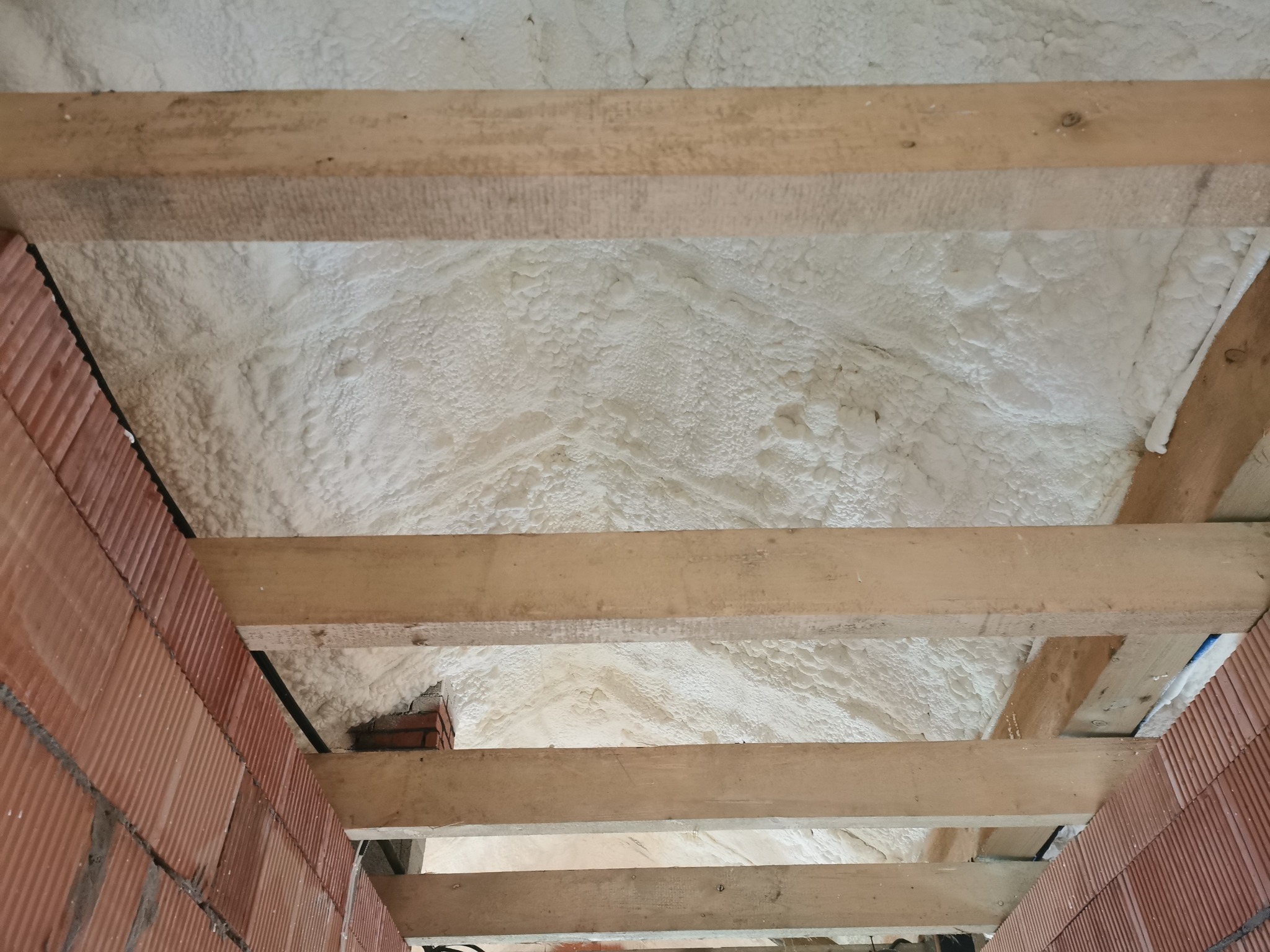 Poddasze ocieplone białą pianką PUR, widoczne drewniane belki konstrukcyjne i fragment muru z czerwonej cegły dziurawki.