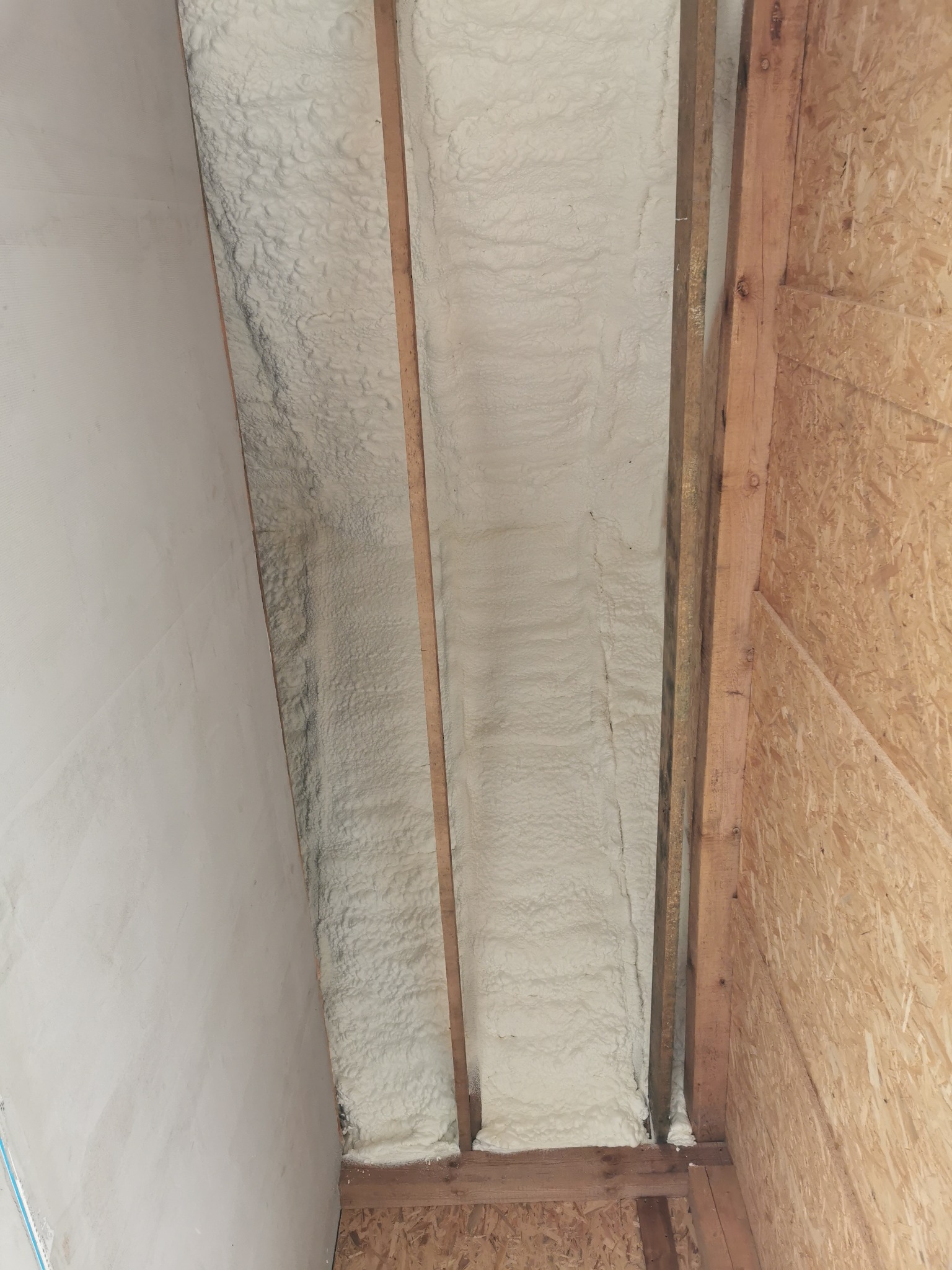 Skośny fragment poddasza z widoczną białą, rozprężoną izolacją natryskową między drewnianymi krokwiami, przylegającą do ściany z płyt OSB i fragmentu otynkowanej ściany.