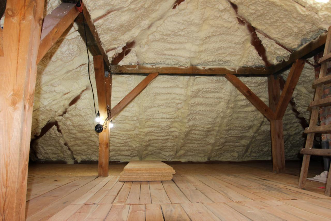 Poddasze z drewnianą konstrukcją i izolacją natryskową, widoczna lampa robocza i stos płyt OSB na drewnianej podłodze.